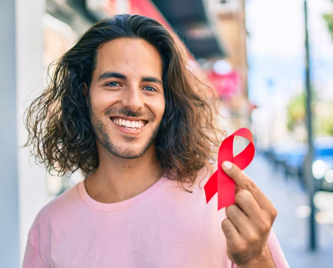 Junger, lateinamerikanischer Mann steht lächelnd und glücklich in der Stadt und hält rote Schleife als Zeichen für HIV-Bewusstsein in der Hand.