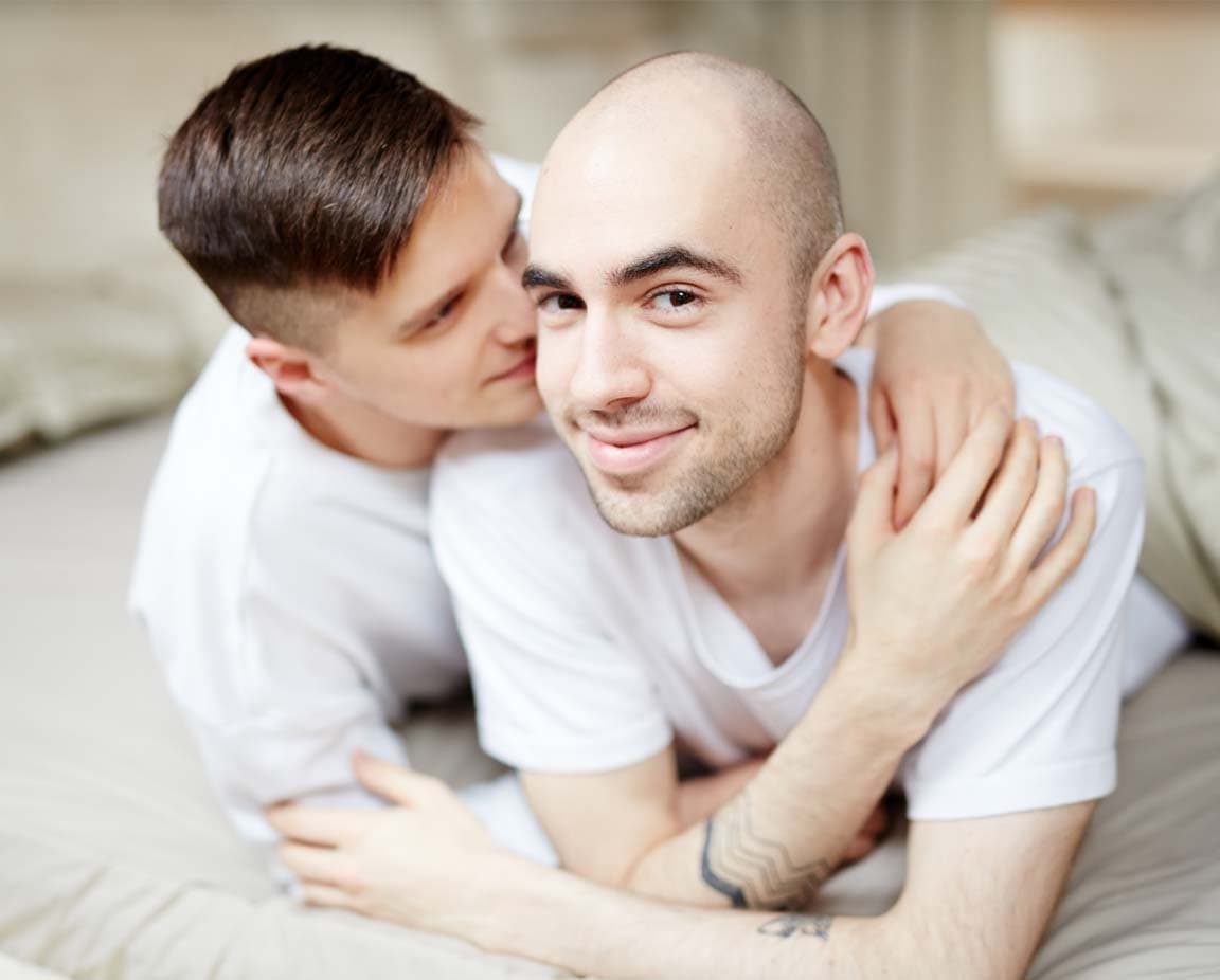 Zärtliches schwules Paar liegt eng umschlungen auf einem Bett. Beide Männer tragen weiße T-Shirts; einer blickt lächelnd in die Kamera, während der andere ihn liebevoll von hinten umarmt. Das Bild strahlt Nähe, Vertrauen und Intimität aus.