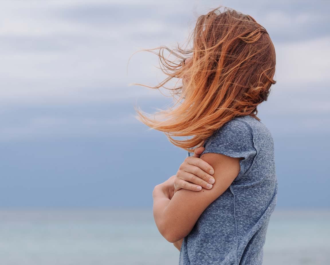 Eine Frau steht allein am Meer mit verschränkten Armen und vom Wind verwehtem Haar. Ihre Haltung wirkt verletzlich und introspektiv – sinnbildlich für Isolation, emotionale Belastung und den inneren Kampf im Kontext einer Suchterkrankung.