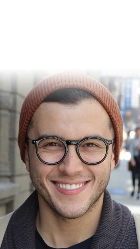 Ein junger Mann mit Brille und Mütze lächelt in die Kamera. Er steht im Freien auf einer städtischen Straße, die im Hintergrund leicht verschwommen ist. Das Bild vermittelt Offenheit, Zugänglichkeit und urbane Lebensrealität – ideal, um jüngere Zielgruppen in der Gesundheitskommunikation direkt anzusprechen.