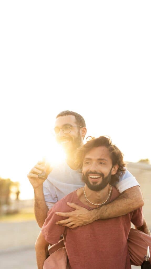 Ein schwules Paar genießt gemeinsam einen sonnigen Moment im Freien. Der eine trägt den anderen lachend auf dem Rücken – Ausdruck von Nähe, Lebensfreude und gegenseitigem Vertrauen.