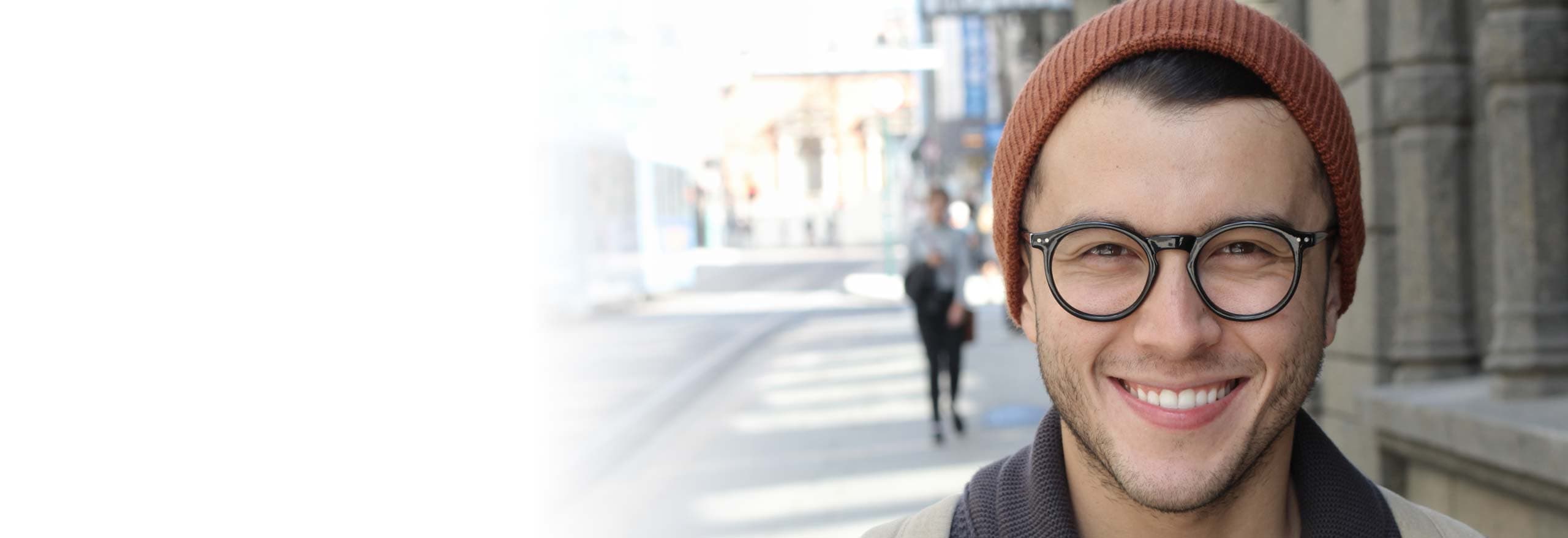 Ein junger Mann mit Brille und Mütze lächelt in die Kamera. Er steht im Freien auf einer städtischen Straße, die im Hintergrund leicht verschwommen ist. Das Bild vermittelt Offenheit, Zugänglichkeit und urbane Lebensrealität – ideal, um jüngere Zielgruppen in der Gesundheitskommunikation direkt anzusprechen.